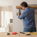 Bricoleur installant une étagère murale avec une perceuse, lunettes de sécurité et niveau à bulle, dans une pièce en rénovation.