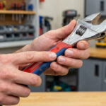 Main d'un bricoleur tenant une clé à molette à manche rouge et bleu, dans un atelier avec des outils en arrière-plan.