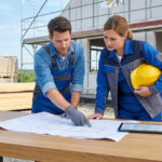 Un homme et une femme examinent des plans sur un chantier. Elle tient un casque, lui pointe les plans. Bâtiment et matériaux en fond.