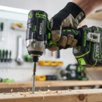 Une main gantée utilise une visseuse à chocs Parkside verte et noire pour visser une vis dans une planche de bois, projetant des copeaux. Arrière-plan flou d'atelier.