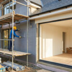 Un ouvrier en casque jaune pose l'isolation extérieure sur une maison neuve en construction, avec échafaudage et matériaux.