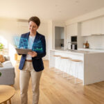 Femme dans un intérieur moderne regardant un plan immobilier et des graphiques financiers projetés depuis sa tablette.