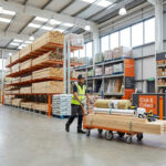 Magasin de bricolage spacieux avec rayonnages de bois, sacs de matériaux, un employé poussant un chariot, et un comptoir Click & Collect.