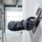 Outillage EDMA : la performance durable pour vos chantiers Main gantée utilisant une truelle pour appliquer de l'enduit sur un mur. Arrière-plan flou d'un chantier en cours.