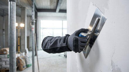 Main gantée utilisant une truelle pour appliquer de l'enduit sur un mur. Arrière-plan flou d'un chantier en cours.