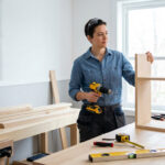 Femme bricoleuse avec lunettes de protection, assemblant un meuble en bois avec une perceuse sur un établi rempli d'outils.