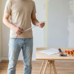 Un homme tient un mètre ruban dans une pièce en rénovation lumineuse. Sur une table, des outils de bricolage et un plan.