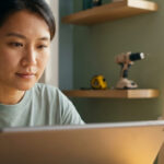 Une femme asiatique utilise une tablette, planifiant un projet de bricolage avec des outils (perceuse, mètre) et du bois visibles en arrière-plan.