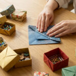 Mains pliant du papier origami bleu pour créer une boîte, entourées de boîtes colorées remplies de petits objets sur une table en bois.
