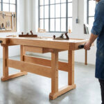 Artisan devant un établi bois stable, avec des outils (rabots, ciseaux) et un tableau mural d'outils, dans un atelier lumineux.