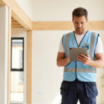 Professionnel en gilet bleu consultant une tablette dans un intérieur moderne. Il représente l'inspection et le contrôle qualité Qualibat.