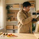 Un homme installe une étagère murale en bois avec une perceuse, portant lunettes et gants. Des outils sont sur le plan de travail.