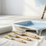 Matériel de peinture neuf sur bâche: rouleau, trois pinceaux, bac, pot de peinture bleue ouvert. Échelle et mur en arrière-plan.