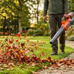 Personne utilisant un souffleur de feuilles orange et gris pour dégager des feuilles d'automne sur une pelouse et un chemin de jardin.