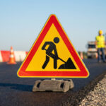 Panneau triangulaire jaune "Travaux" en gros plan sur route neuve avec ouvriers et obstacles de chantier en arrière-plan flou.