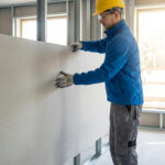 Ouvrier avec casque et gants installant une grande plaque de Fermacell sur une ossature métallique dans un bâtiment en construction.