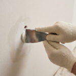 Mains gantées appliquant de l'enduit de rebouchage avec une spatule pour réparer un trou dans un mur clair. Outils en arrière-plan.