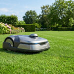 Robot tondeuse autonome gris sur pelouse luxuriante. En arrière-plan, des hortensias colorés et une haie bien taillée.