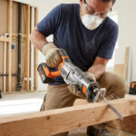 Homme masqué avec lunettes et gants coupe une poutre en bois avec une scie sabre sans fil sur un chantier de rénovation.