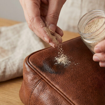 Une main saupoudre de la terre de Sommières sur une tache sombre sur un sac en cuir marron.