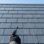 Main gantée appliquant un hydrofuge sur une toiture en tuiles grises. Des gouttes d'eau perlent sur la surface traitée.